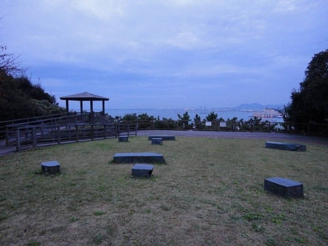 芝生の広場を見下ろす写真。手前中央に六つの大・小さまざまな形の低い石製ベンチが円を描くように点在し、左奥に木製のあずまやとそれにつながるウッドデッキの通路が見える。中景には手すりのある柵越しに海面が広がり、波打ち際には緑の木立が並ぶ。